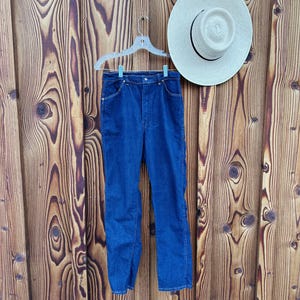 May include: A pair of blue denim jeans hanging on a wooden hanger against a wood panel background. The jeans have a classic straight leg style and a button fly.