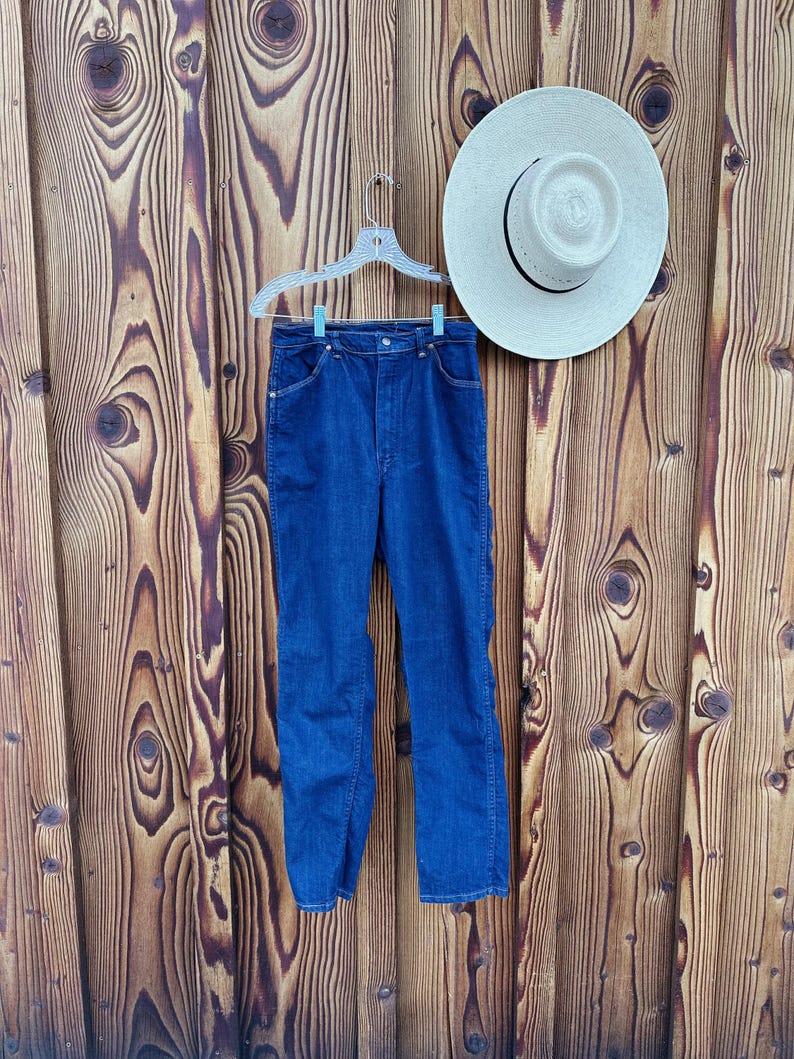 May include: A pair of blue denim jeans hanging on a wooden hanger against a wood panel background. The jeans have a classic straight leg style and a button fly.