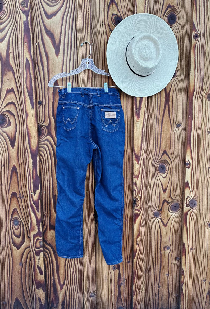 May include: A pair of dark blue denim jeans hanging on a white hanger against a woodgrain background. The jeans have a classic five-pocket style and a leather patch on the back right pocket with the Wrangler logo.