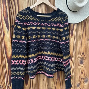May include: A black sweater with a colorful geometric pattern in pink, gold, and gray. The sweater is hanging on a wooden hanger against a wooden background. A straw hat is visible in the background.