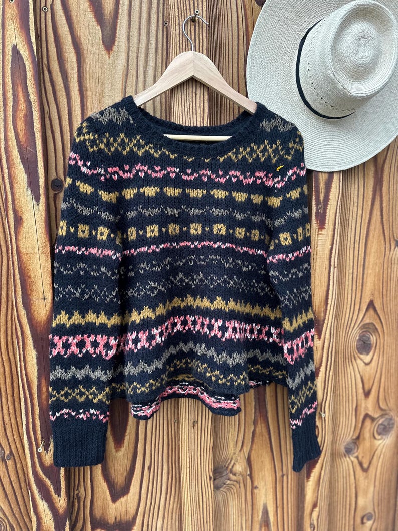 May include: A black sweater with a colorful geometric pattern in pink, gold, and gray. The sweater is hanging on a wooden hanger against a wooden background. A straw hat is visible in the background.