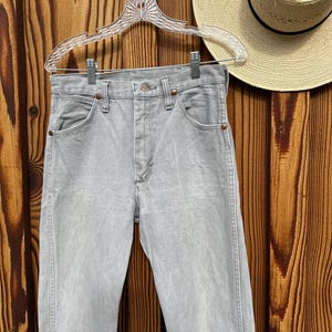Pu&ograve; includere: Jeans di denim grigio chiaro appesi a una gruccia trasparente su uno sfondo di legno. I jeans hanno un classico design a cinque tasche e una chiusura con bottone e cerniera. Un cappello di paglia &egrave; visibile sullo sfondo.