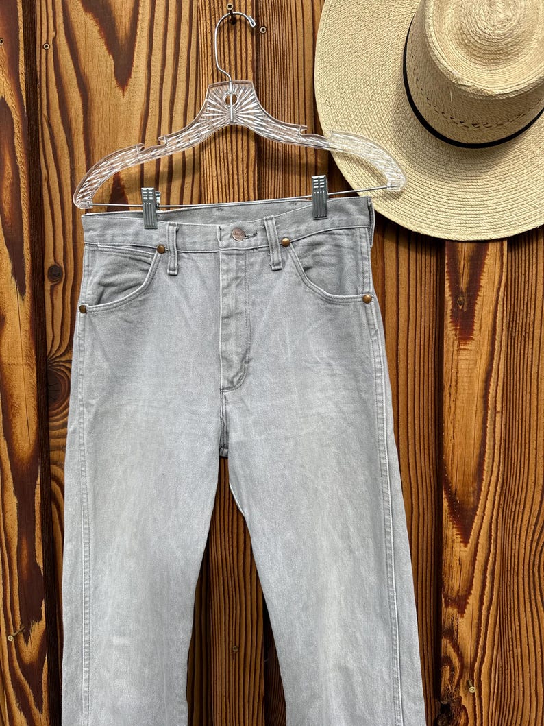 Pu&ograve; includere: Jeans di denim grigio chiaro appesi a una gruccia trasparente su uno sfondo di legno. I jeans hanno un classico design a cinque tasche e una chiusura con bottone e cerniera. Un cappello di paglia &egrave; visibile sullo sfondo.