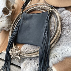 Pode incluir: Uma bolsa de couro preta com detalhes longos de franjas. A bolsa est&aacute; sobre uma superf&iacute;cie de pele marrom e branca com uma corda e um chap&eacute;u de cowboy.