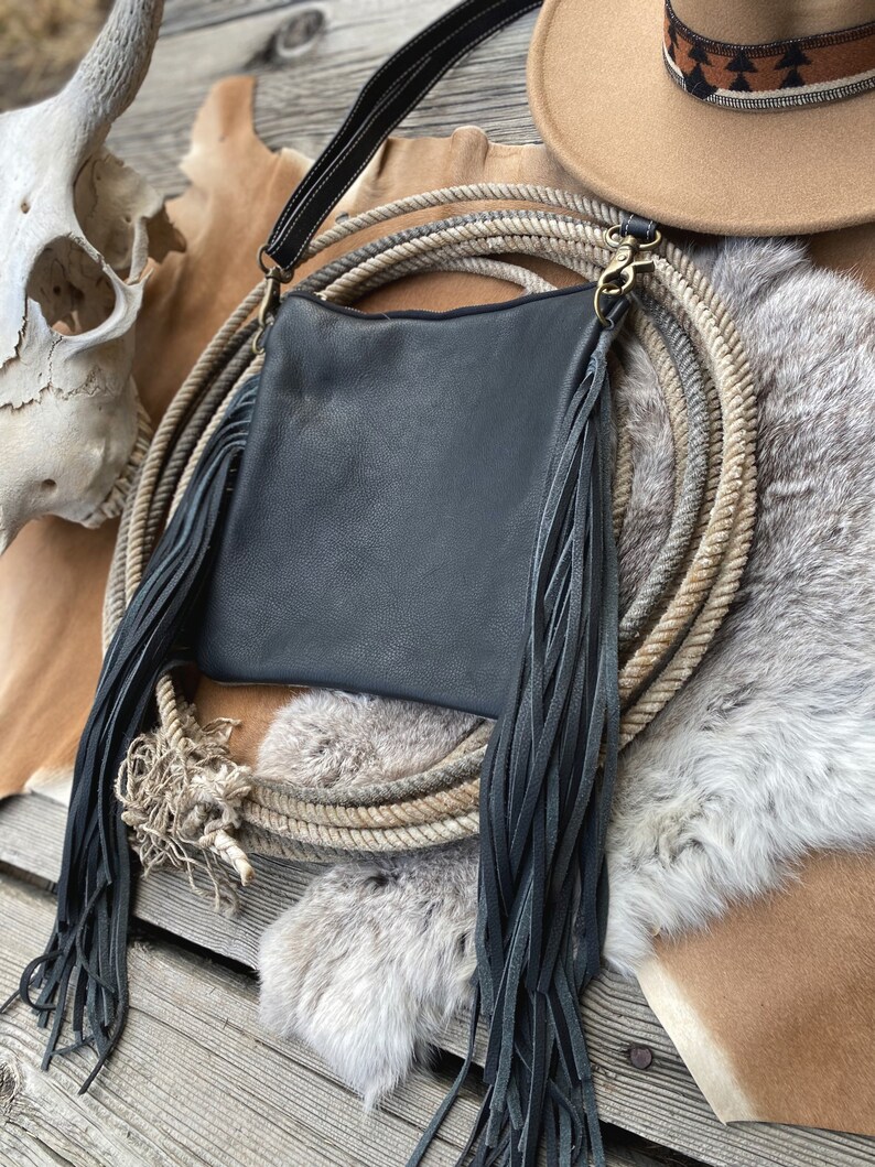 Pode incluir: Uma bolsa de couro preta com detalhes longos de franjas. A bolsa est&aacute; sobre uma superf&iacute;cie de pele marrom e branca com uma corda e um chap&eacute;u de cowboy.