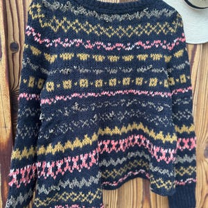 May include: A navy blue knit sweater with a geometric pattern in pink, gold, and gray. The sweater is hanging on a wooden hanger against a wooden background. A white hat with a black band is visible in the background.