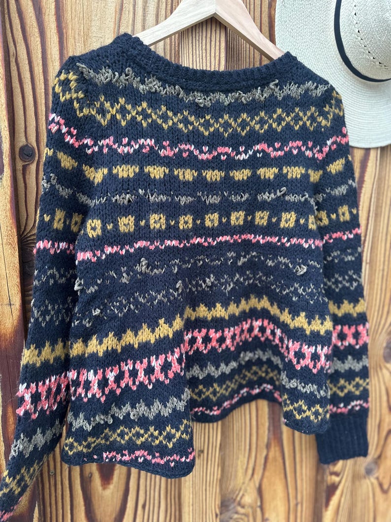 May include: A navy blue knit sweater with a geometric pattern in pink, gold, and gray. The sweater is hanging on a wooden hanger against a wooden background. A white hat with a black band is visible in the background.