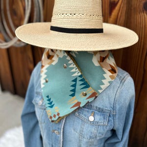 May include: A wide-brimmed straw hat with a black band, worn over a blue denim jacket with a turquoise and brown patterned scarf. The scarf has a geometric design and is secured with metal snaps.