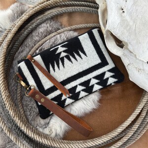 Puede incluir: Un clutch con estampado geométrico en blanco y negro con una correa de cuero marrón y un cierre plateado. El clutch está sobre una superficie de piel marrón y blanca.