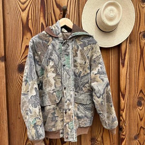 May include: A camouflage jacket with a hood and two front pockets hangs on a wooden hanger. The jacket features a brown hood and cuffs, with a button-up front. A straw hat with a black band hangs on the wooden wall.