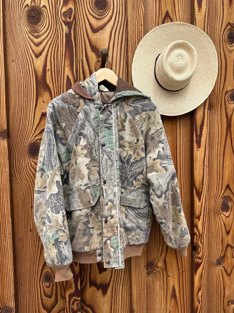 May include: A camouflage jacket with a hood and two front pockets hangs on a wooden hanger. The jacket features a brown hood and cuffs, with a button-up front. A straw hat with a black band hangs on the wooden wall.