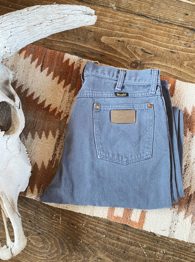 May include: A pair of gray Wrangler jeans with a brown leather patch on the back pocket. The patch has the Wrangler logo in white.