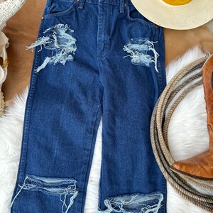 May include: Distressed dark blue denim jeans with multiple rips and frayed edges. The jeans have a high waist and a straight leg.