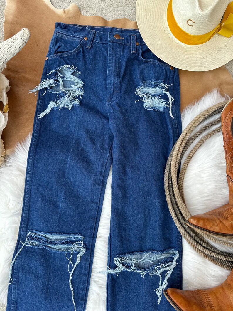 May include: Distressed dark blue denim jeans with multiple rips and frayed edges. The jeans have a high waist and a straight leg.