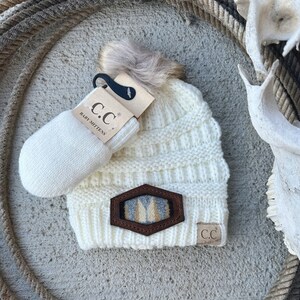 May include: White knit baby beanie with a faux fur pom pom and a brown leather patch with a geometric design. The beanie is paired with a white knit baby mitten that says "C.C. Baby Mittens".