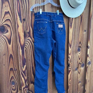 May include: A pair of blue denim jeans with a high waist and a straight leg. The jeans have a brown leather patch on the back right pocket with the Wrangler logo.