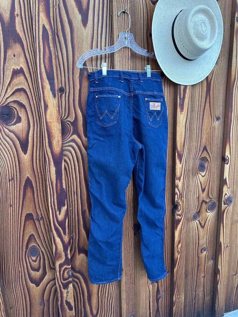 May include: A pair of blue denim jeans with a high waist and a straight leg. The jeans have a brown leather patch on the back right pocket with the Wrangler logo.