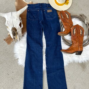 May include: A pair of dark blue denim jeans with a classic straight leg fit. The jeans have a brown leather patch with the Wrangler logo on the back waistband.