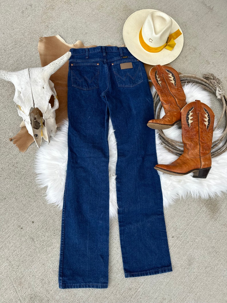 May include: A pair of dark blue denim jeans with a classic straight leg fit. The jeans have a brown leather patch with the Wrangler logo on the back waistband.