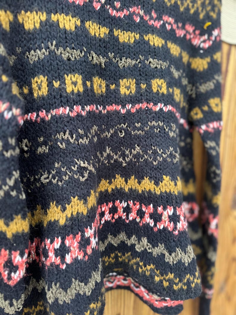 May include: Close-up of a dark blue knit sweater with a geometric pattern. The sweater features repeating patterns in gold, pink, and white. The knitwear has a textured appearance, with visible stitches and a cozy aesthetic.
