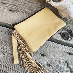 May include: A light yellow leather clutch with a fringe detail. The clutch has a black zipper and is sitting on a wooden surface.