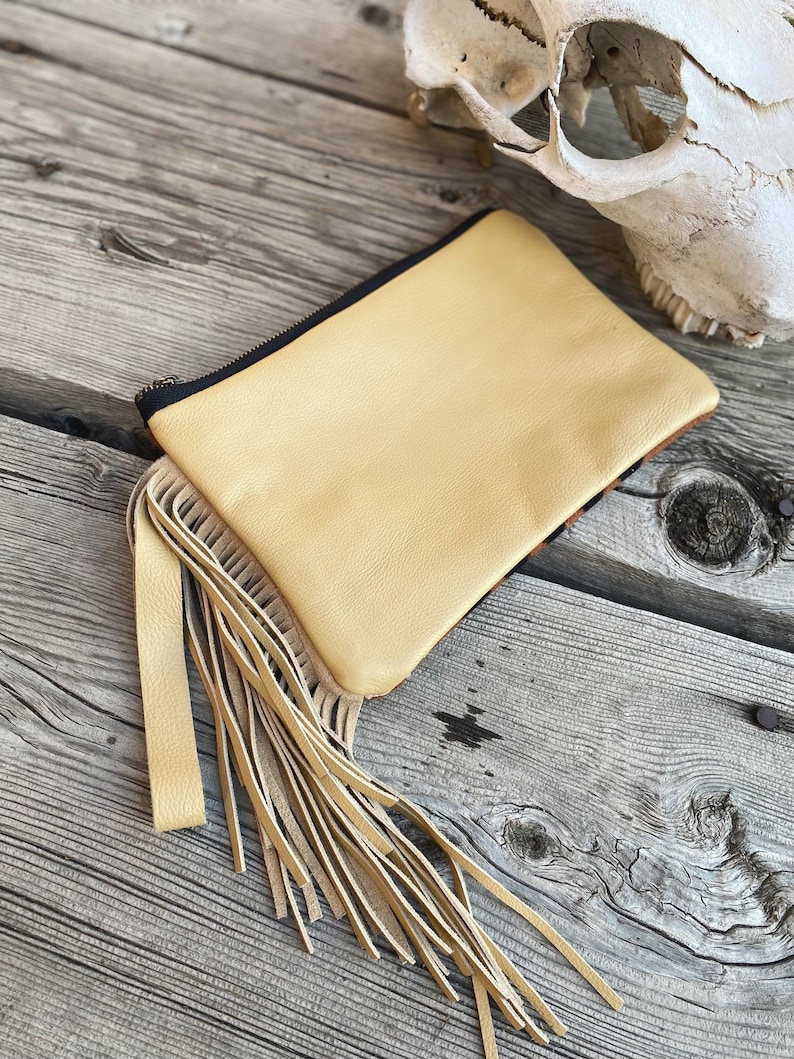 May include: A light yellow leather clutch with a fringe detail. The clutch has a black zipper and is sitting on a wooden surface.
