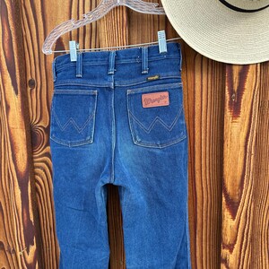 Puede incluir: Un par de pantalones vaqueros de mezclilla azul oscuro con una etiqueta Wrangler de cuero marr&oacute;n en el bolsillo trasero.