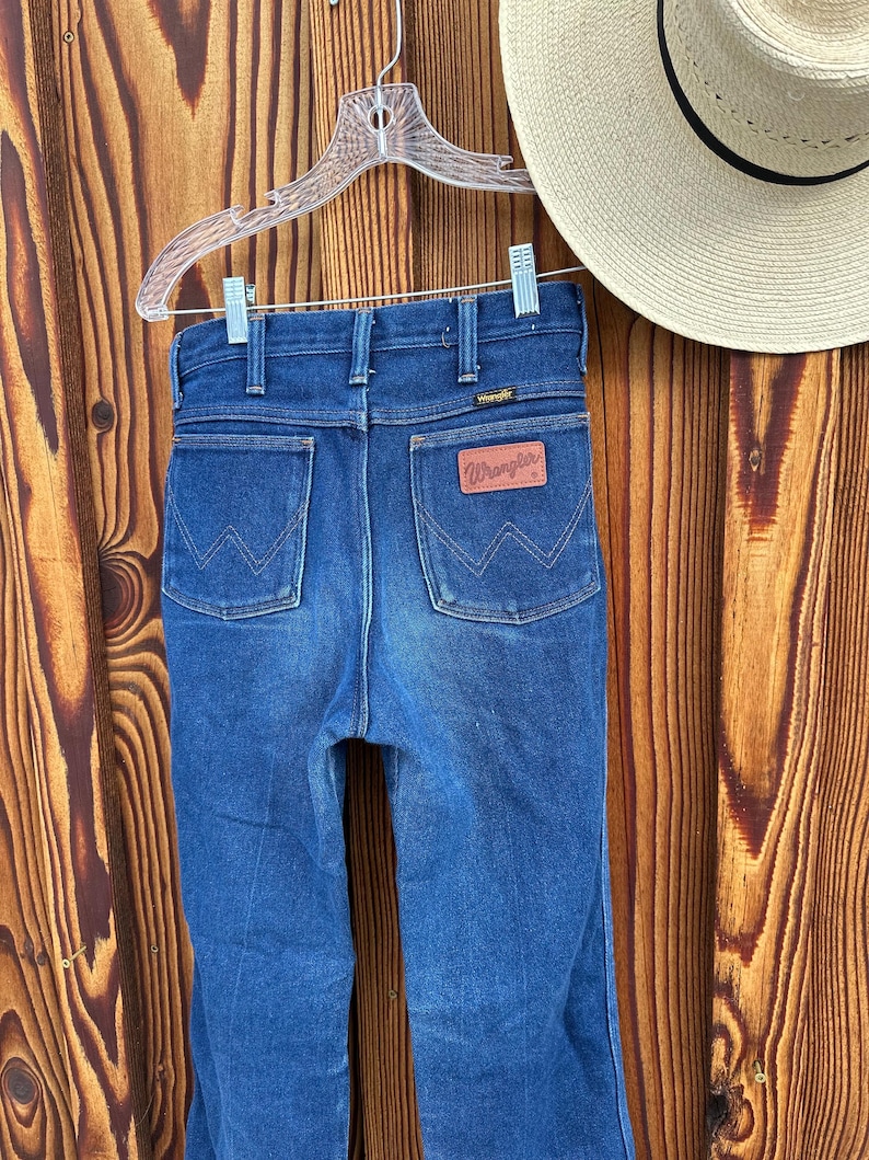Puede incluir: Un par de pantalones vaqueros de mezclilla azul oscuro con una etiqueta Wrangler de cuero marr&oacute;n en el bolsillo trasero.