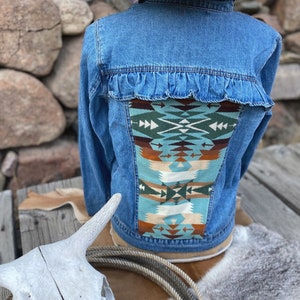 K&ouml;nnte beinhalten: Eine blaue Jeansjacke mit R&uuml;schenbesatz und einem bunten, s&uuml;dwestlichen Muster auf der R&uuml;ckseite. Die Jacke wird von einer Person mit einem Strohhut getragen.