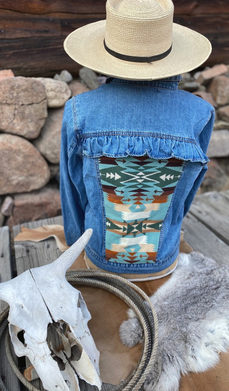 K&ouml;nnte beinhalten: Eine blaue Jeansjacke mit R&uuml;schenbesatz und einem bunten, s&uuml;dwestlichen Muster auf der R&uuml;ckseite. Die Jacke wird von einer Person mit einem Strohhut getragen.