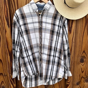 May include: A long-sleeved plaid shirt in white, black, and brown hangs on a wooden hanger. The shirt has a button-down collar and a pocket on the left chest. A straw hat with a black band rests to the right of the shirt.