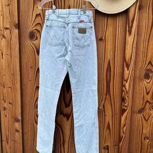 May include: Pale blue denim jeans hanging on a clear hanger. The jeans feature a classic five-pocket design, a brown leather patch, and the brand name "Wrangler" on the back pocket. A straw hat is visible in the background.