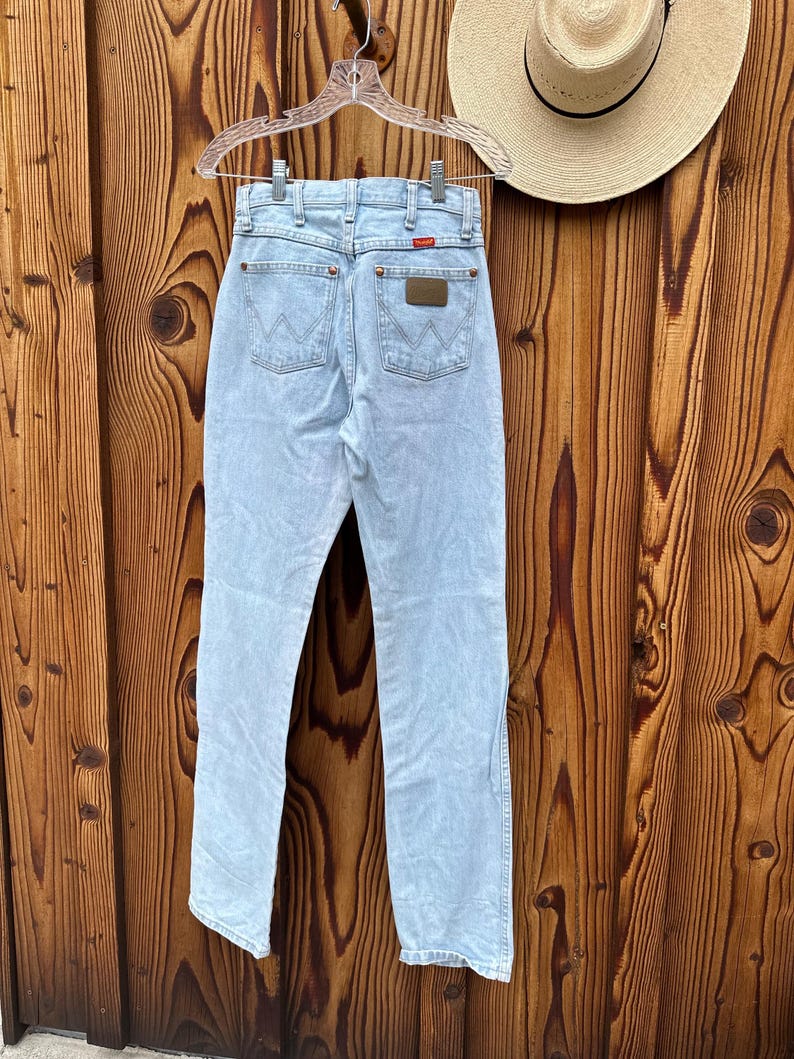May include: Pale blue denim jeans hanging on a clear hanger. The jeans feature a classic five-pocket design, a brown leather patch, and the brand name "Wrangler" on the back pocket. A straw hat is visible in the background.