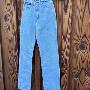 May include: Light blue denim jeans hanging on a clear hanger. The jeans have a classic five-pocket design, a button and zipper closure, and a straight leg. The jeans are made of a light wash denim material.