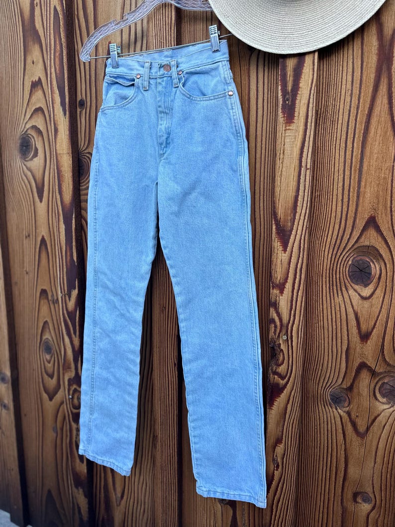 May include: Light blue denim jeans hanging on a clear hanger. The jeans have a classic five-pocket design, a button and zipper closure, and a straight leg. The jeans are made of a light wash denim material.