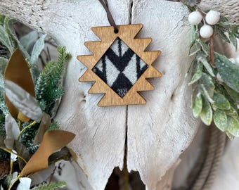 May include: Wooden ornament with a geometric design, featuring a black and white pattern. The ornament is suspended by a brown leather cord. The background includes a white skull, rope, and greenery, creating a rustic, southwestern aesthetic.