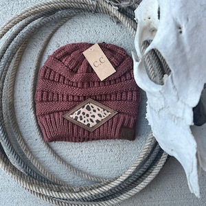 May include: A maroon knit beanie with a brown leather patch featuring a leopard print. The beanie has a small brown tag with the letters "C.C." on it.