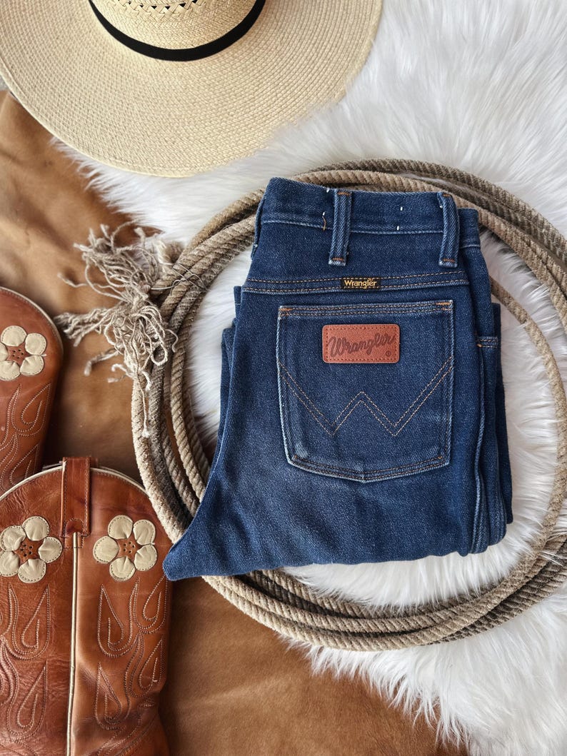 Puede incluir: Un par de pantalones vaqueros de mezclilla azul oscuro con una etiqueta Wrangler de cuero marr&oacute;n. Los pantalones vaqueros est&aacute;n doblados y colocados sobre una superficie blanca y esponjosa con un fondo de cuero marr&oacute;n. Tambi&eacute;n se puede ver un sombrero de vaquero marr&oacute;n y un par de botas de vaquero marrones en la imagen.