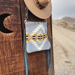 Pode incluir: Uma bolsa crossbody cinza e branca com padrão e detalhes de franjas. A bolsa está pendurada em uma superfície de madeira.
