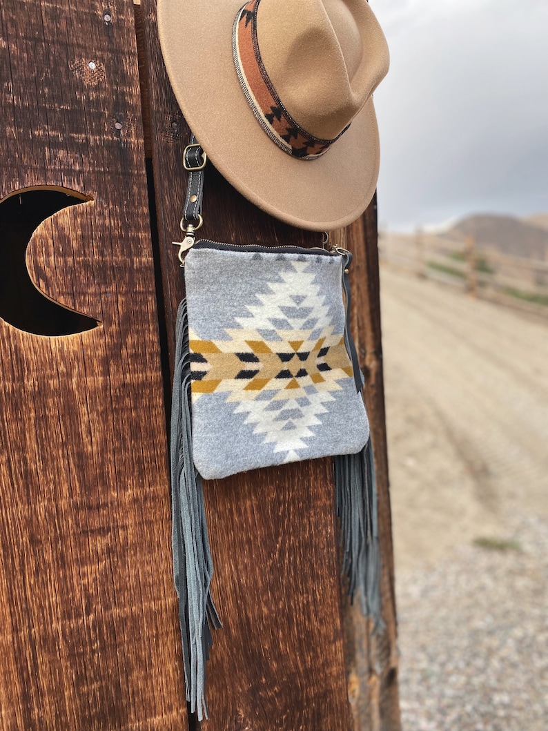 Pode incluir: Uma bolsa crossbody cinza e branca com padr&atilde;o e detalhes de franjas. A bolsa est&aacute; pendurada em uma superf&iacute;cie de madeira.