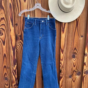 Puede incluir: Un par de pantalones vaqueros azules de pierna ancha, colgados de una percha de pl&aacute;stico blanca sobre un fondo de madera.