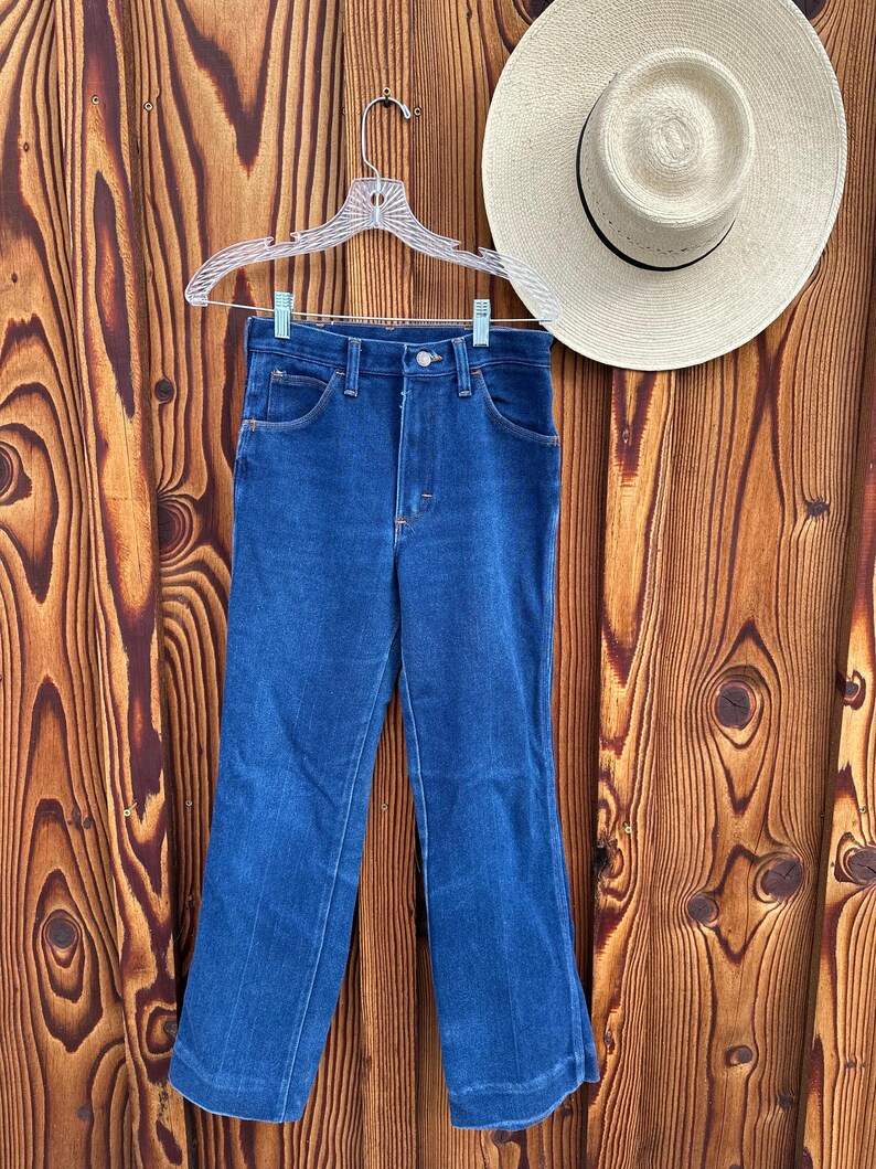 Puede incluir: Un par de pantalones vaqueros azules de pierna ancha, colgados de una percha de pl&aacute;stico blanca sobre un fondo de madera.