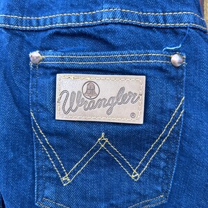 May include: A close-up of a blue denim back pocket with a brown leather Wrangler patch that says "Blue Bell" and yellow stitching detail.
