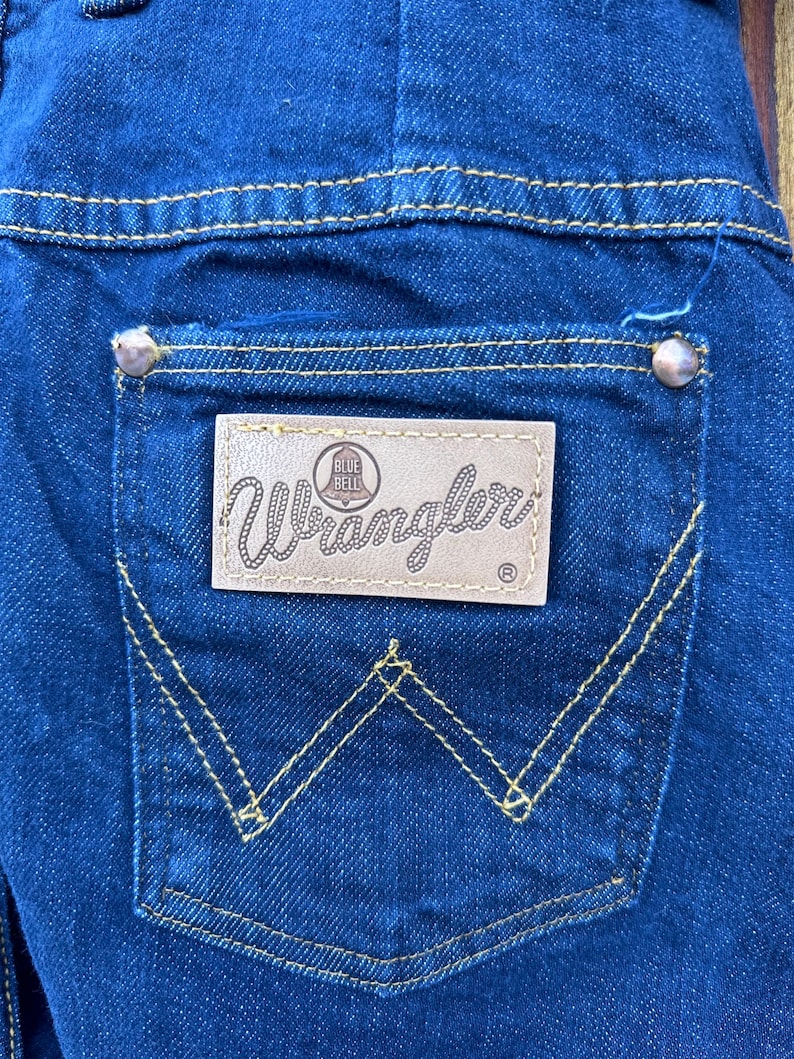 May include: A close-up of a blue denim back pocket with a brown leather Wrangler patch that says "Blue Bell" and yellow stitching detail.