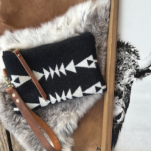 May include: A black and white geometric patterned clutch with a brown leather strap. The clutch is lying on a brown and white fur surface.