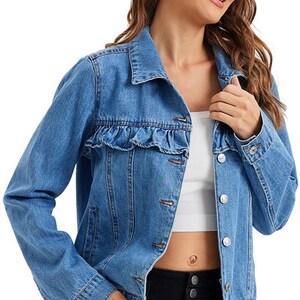 K&ouml;nnte beinhalten: Eine hellblaue Jeansjacke mit R&uuml;schenbesatz an der Brust. Die Jacke hat einen Knopfverschluss und lange &Auml;rmel.