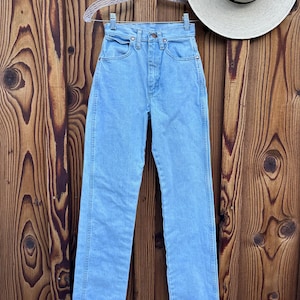 May include: Light blue denim jeans hanging on a clear hanger. The jeans have a classic five-pocket design and a straight leg. A straw hat with a black band is visible in the background. The jeans are displayed against a wooden background.
