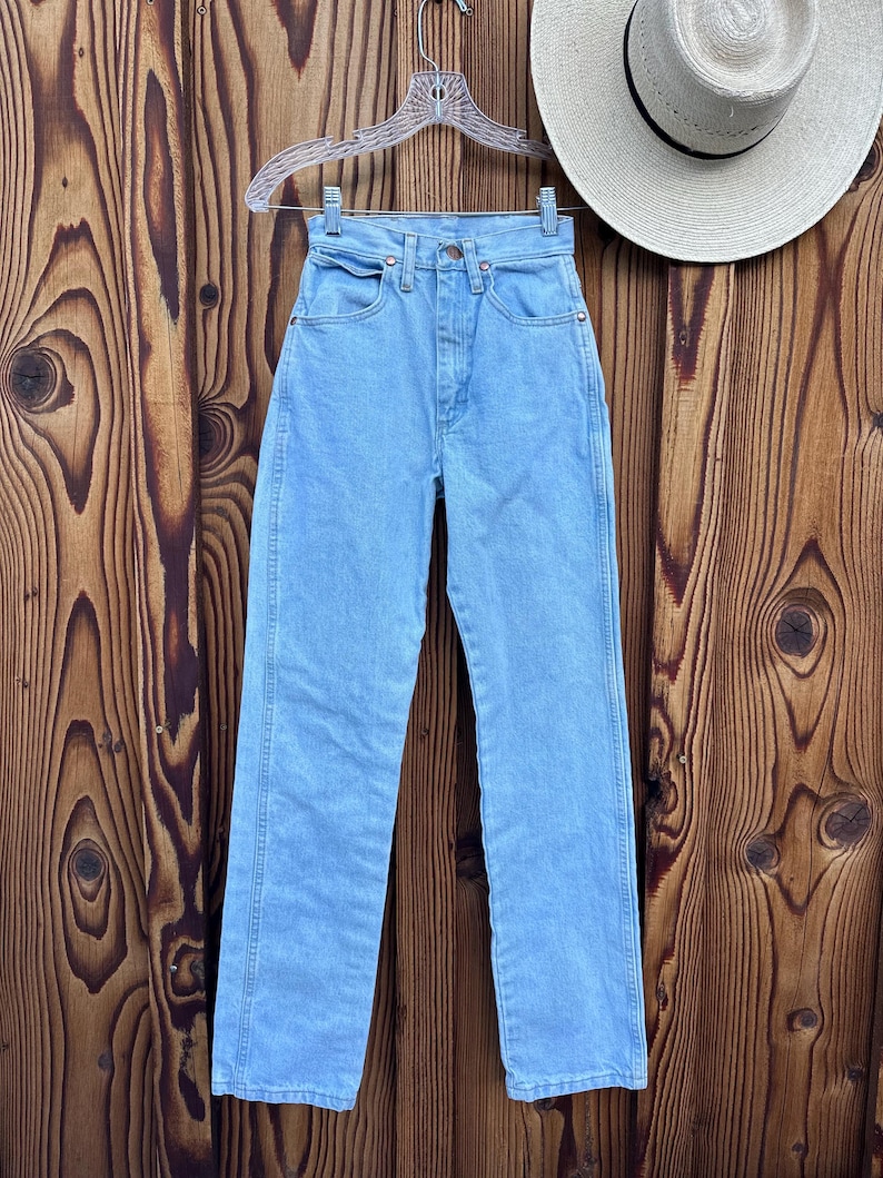 May include: Light blue denim jeans hanging on a clear hanger. The jeans have a classic five-pocket design and a straight leg. A straw hat with a black band is visible in the background. The jeans are displayed against a wooden background.