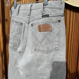 Pu&ograve; includere: Jeans Wrangler grigio chiaro appesi a una gruccia trasparente. I jeans presentano una toppa in pelle marrone con il logo Wrangler sulla tasca posteriore. I jeans sono in denim e hanno un classico design a cinque tasche.