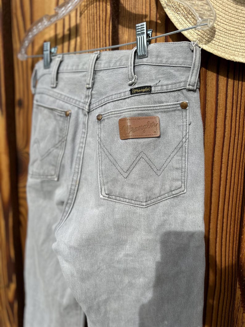 Pu&ograve; includere: Jeans Wrangler grigio chiaro appesi a una gruccia trasparente. I jeans presentano una toppa in pelle marrone con il logo Wrangler sulla tasca posteriore. I jeans sono in denim e hanno un classico design a cinque tasche.